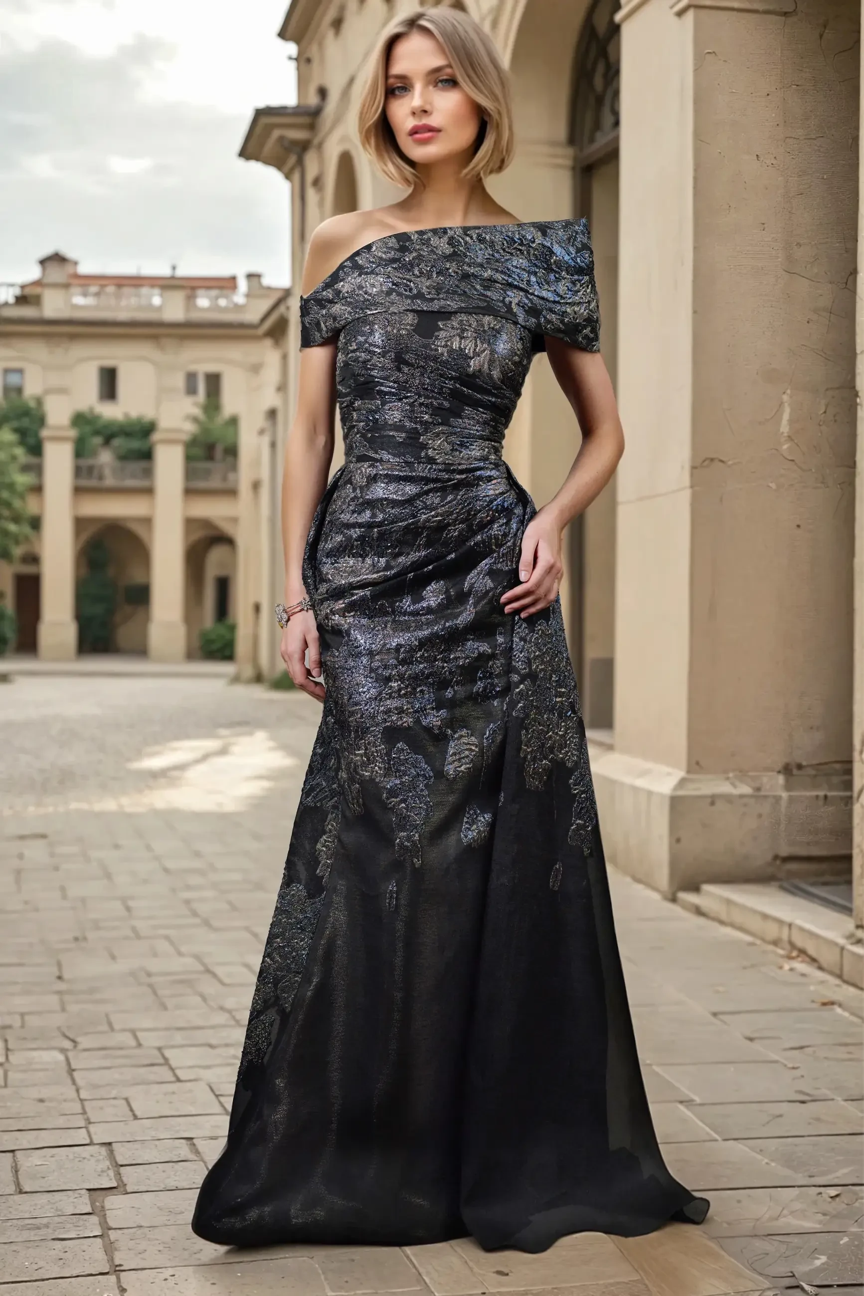 A woman in an elegant off-shoulder black gown with intricate patterns, standing in a stone courtyard surrounded by architectural features.
