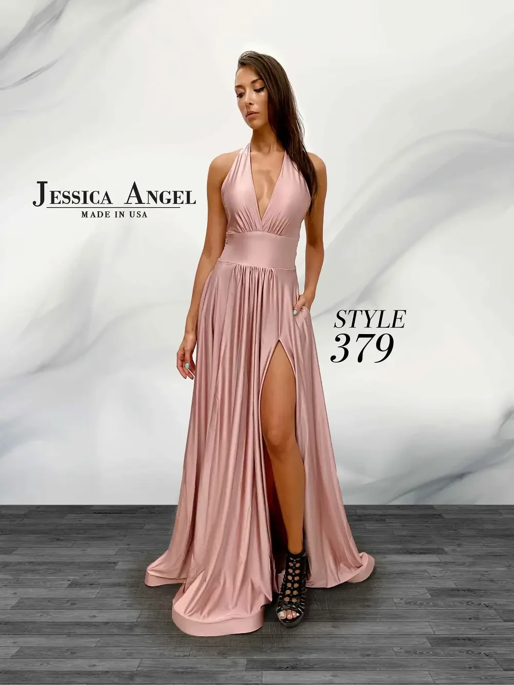 A woman wearing a long, blush pink dress with a deep V-neck and a thigh-high slit, standing against a marble-like background. The dress is labeled as 
