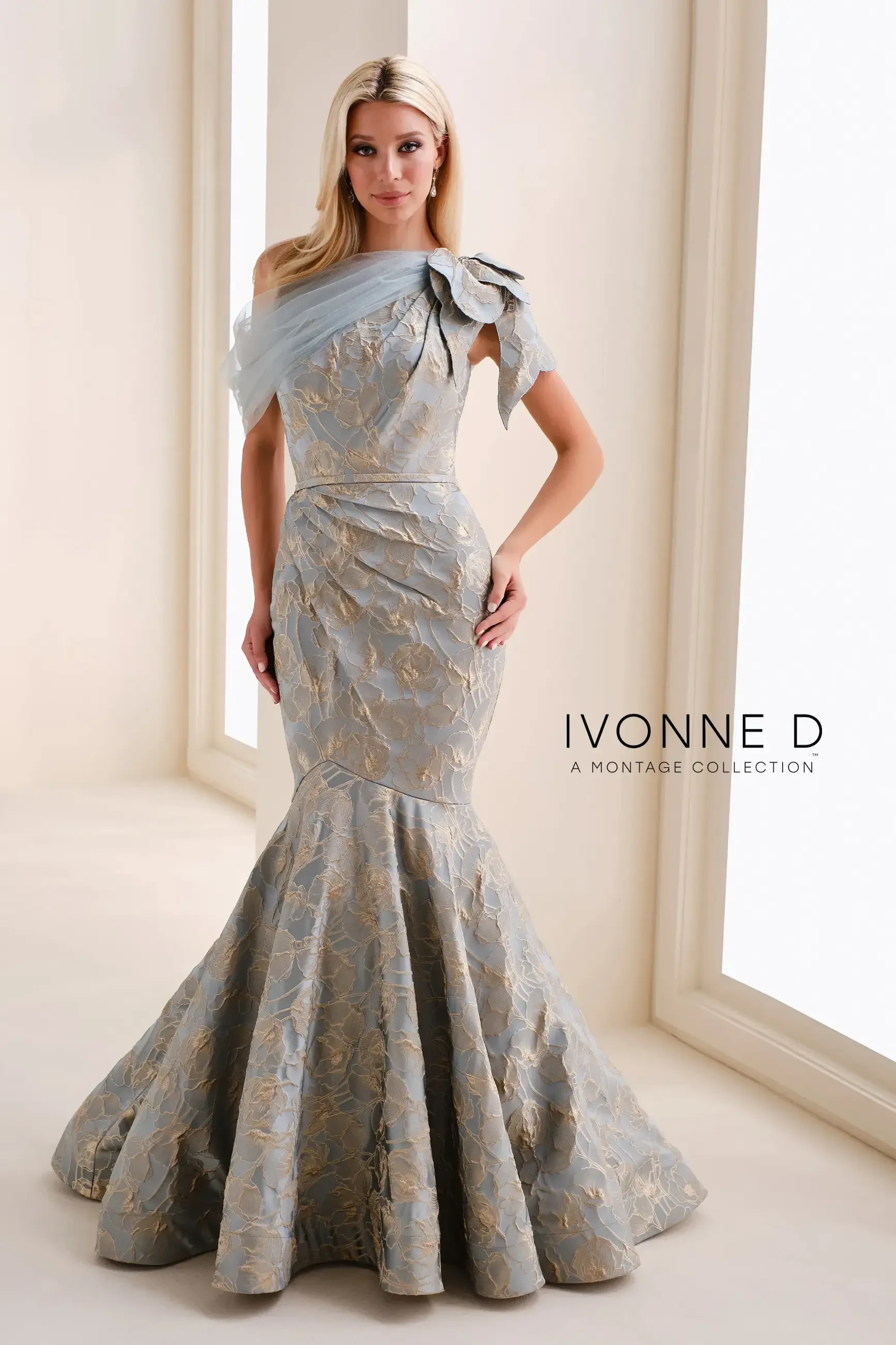A woman wearing a light gray formal gown with a mermaid silhouette and floral accents, standing in an elegant setting. The gown features intricate patterns and a fitted bodice.